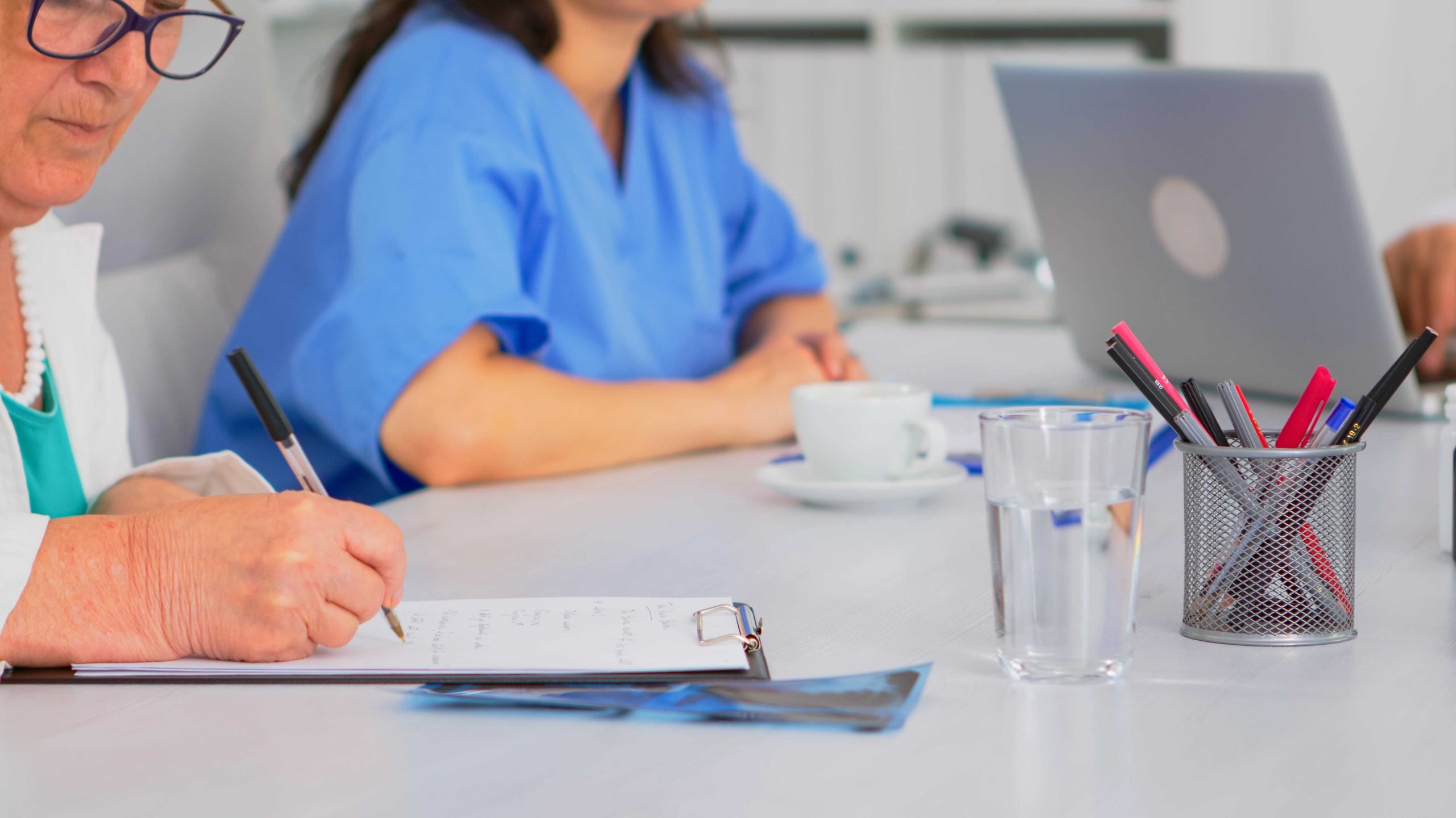 compressed close up elderly woman doctor taking notes clipboard while coworkers discussing background writing laptop brainstorming profesional teamworker having medical meeting scaled
